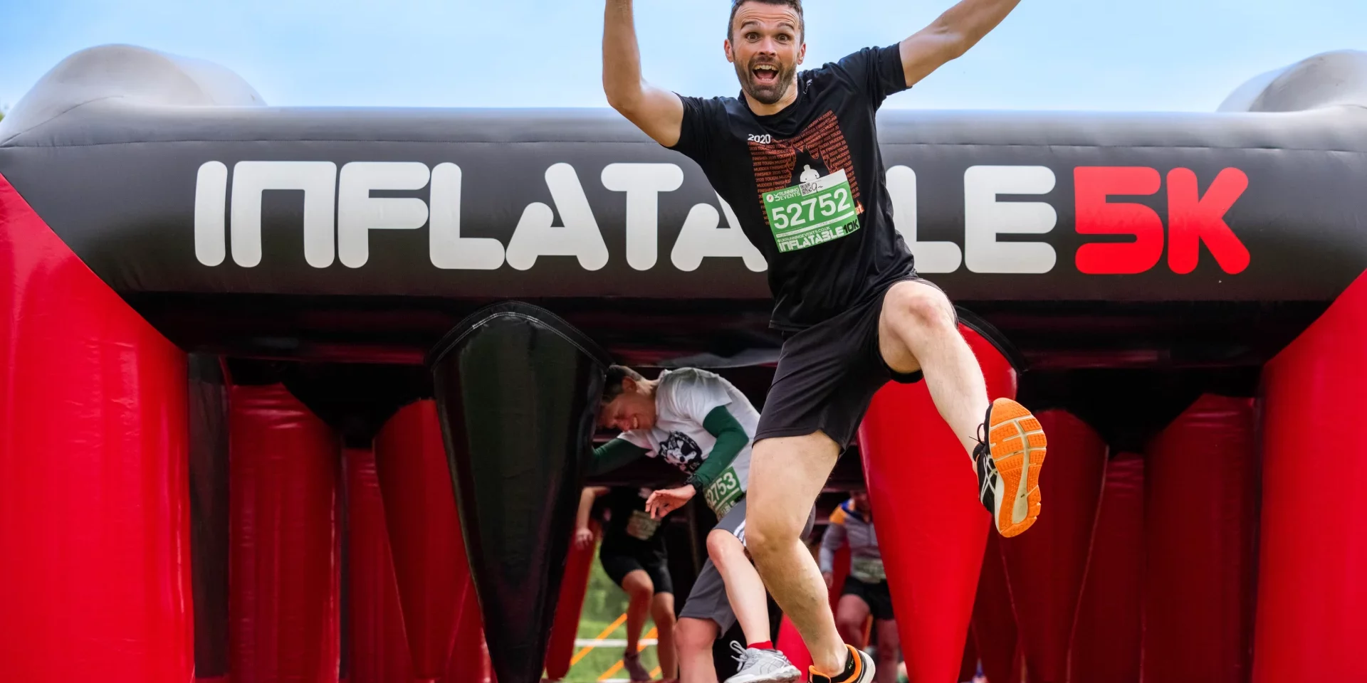 A man joyfully jumps above an inflatable archway at the finish line of a 5K run.